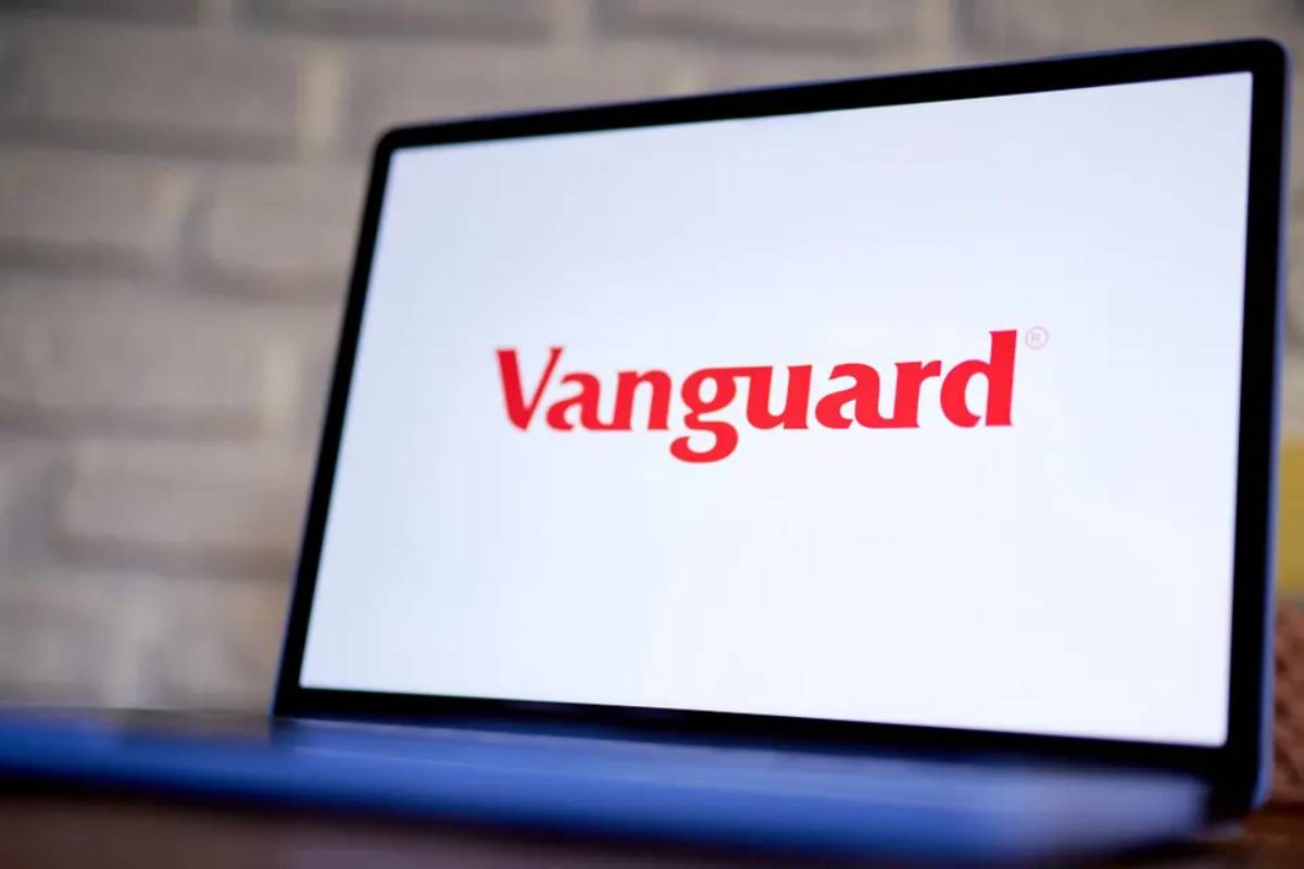 A close-up of a laptop screen displaying the red Vanguard logo on a white background, with a blurred brick wall behind it.