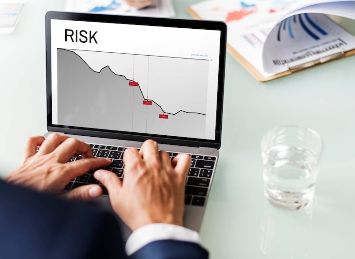 A person in a suit types on a laptop displaying a downward trend graph titled RISK, with a glass of water nearby.