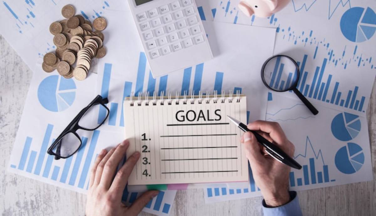 A person writes GOALS in a notebook, surrounded by charts, coins, a calculator, glasses, and a magnifying glass, symbolizing financial planning.