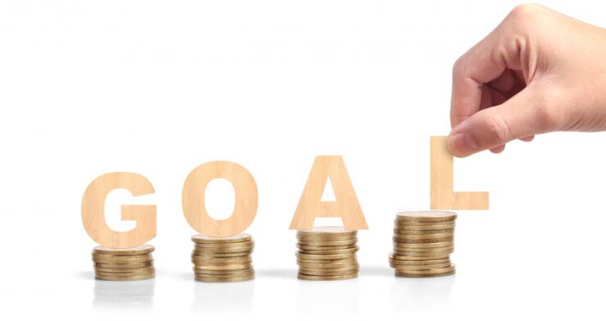 A hand places the letter L atop stacks of coins that spell GOAL, symbolizing financial achievement and aspirations.