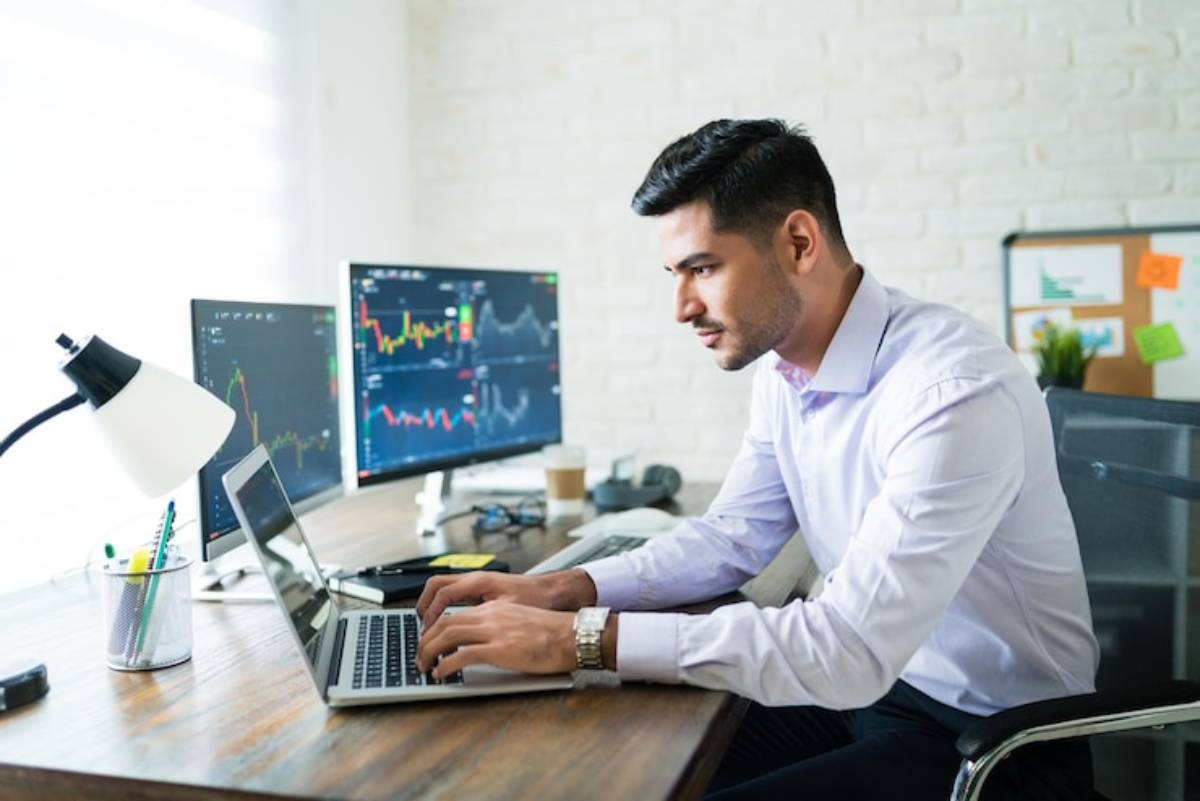 A man in a light purple shirt works intently on a laptop at a desk, surrounded by computer screens displaying financial data.
