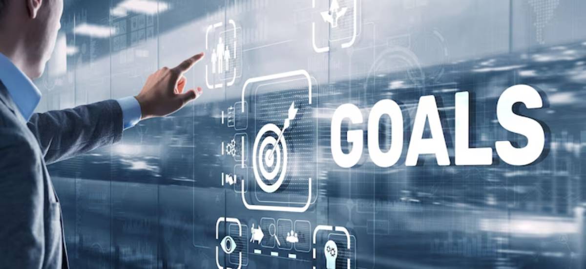 A businessman points at a digital display highlighting the word GOALS, surrounded by various icons representing data and teamwork.