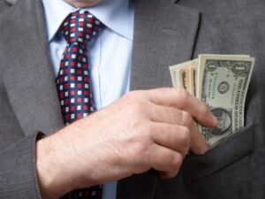 A businessman in a gray suit discreetly places dollar bills into his suit pocket, emphasizing themes of money and secrecy.