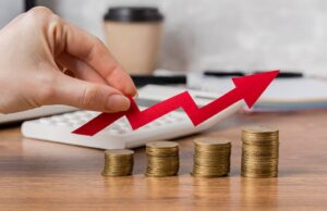 A hand places a red arrow graph above stacked coins, symbolizing financial growth and investment success on a wooden desk.