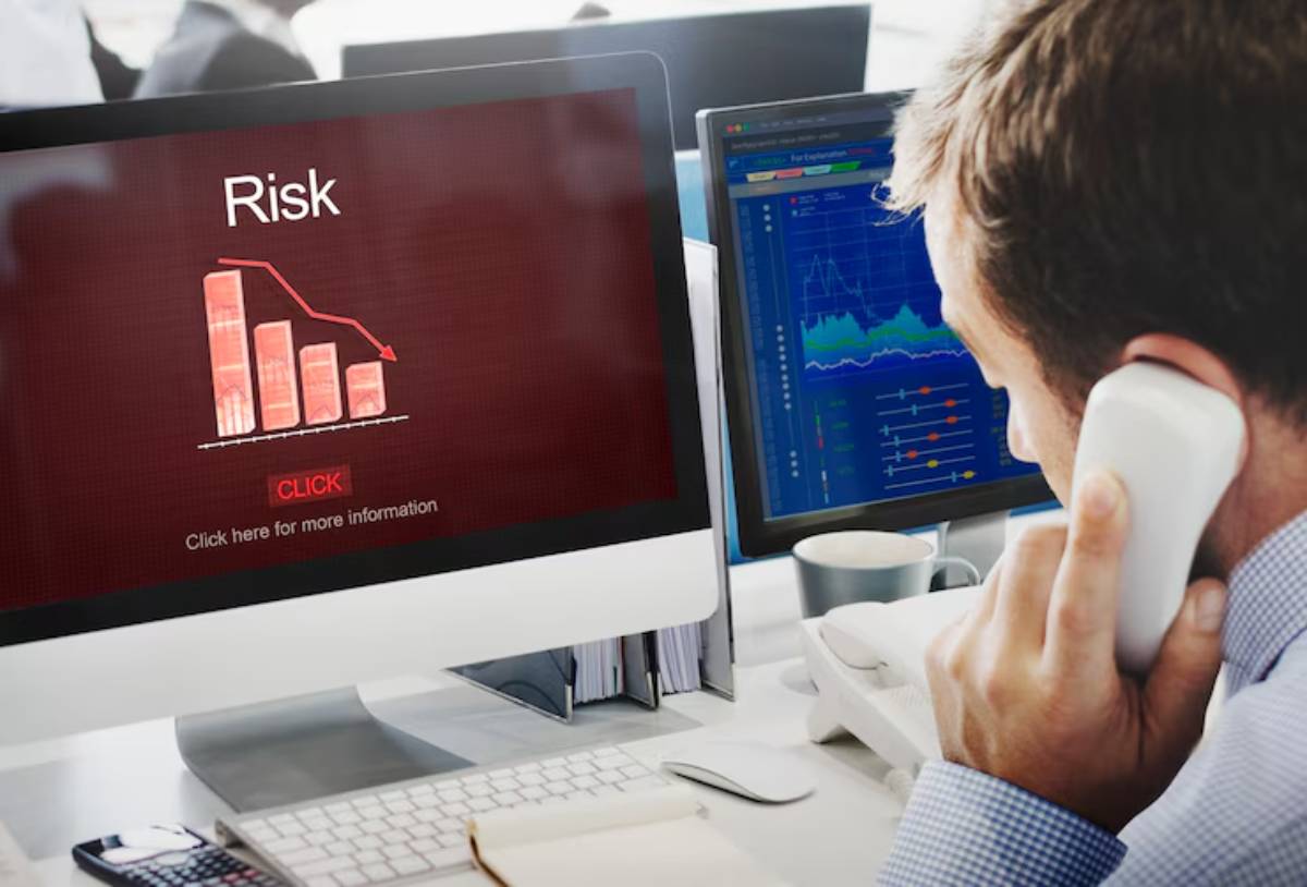 A man on the phone views a computer screen displaying a declining risk graph, emphasizing financial analysis in a busy office setting.