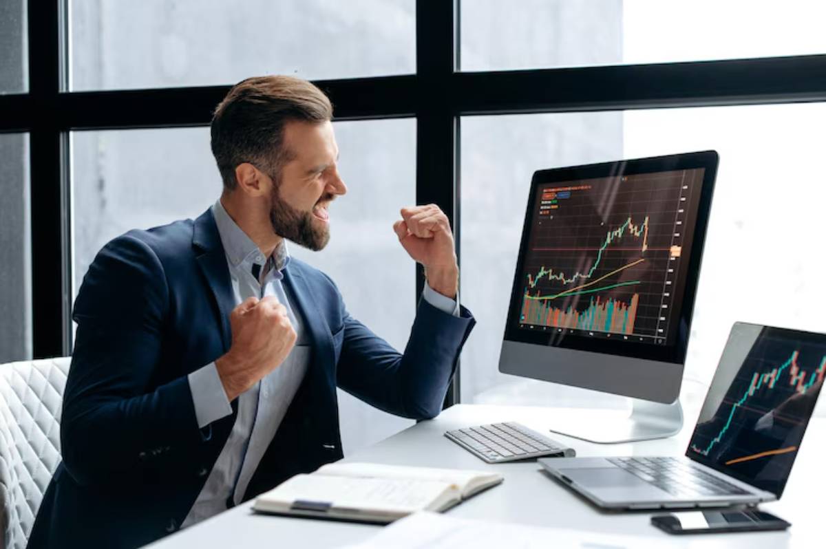 A businessman celebrates a successful investment outcome while analyzing stock market graphs on his computer screens.