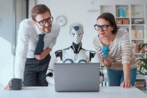 Two colleagues observe a humanoid robot at a desk with a laptop, while one holds a cup and the other leans in for a closer look.