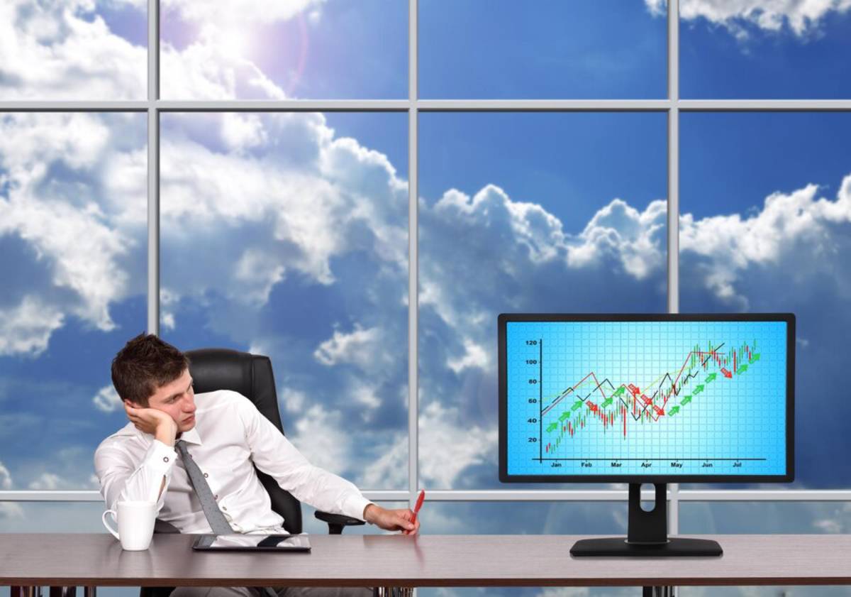 A man in formal attire sits at a desk, looking pensively at a computer screen displaying a colorful financial graph, with clouds outside the window.