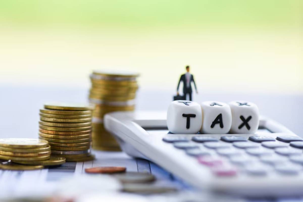 A miniature businessman stands on a calculator beside stacked coins, with dice displaying the word TAX. Financial theme.