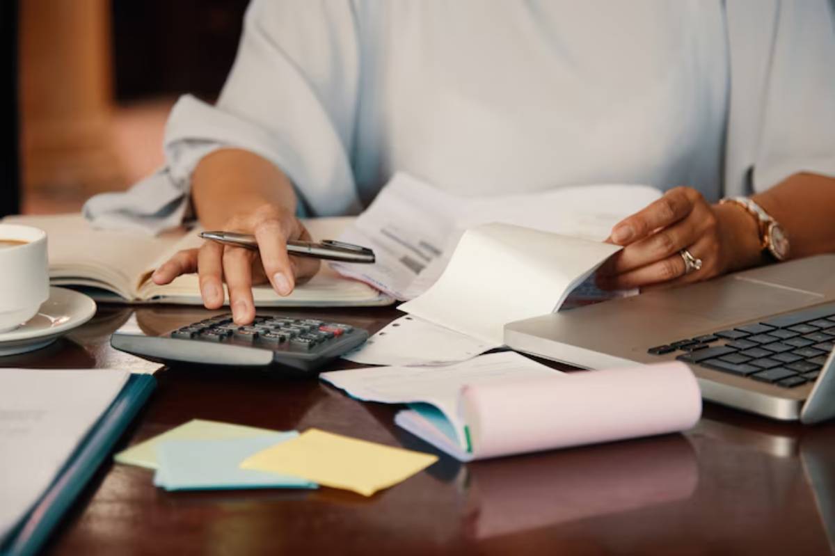 A person calculates expenses with a calculator, surrounded by paperwork, a laptop, sticky notes, and a cup of coffee.