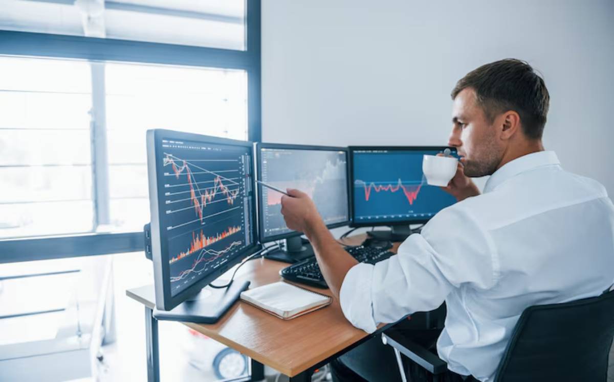 A professional man analyzes data on multiple computer screens, sipping coffee in a modern office setting with large windows.
