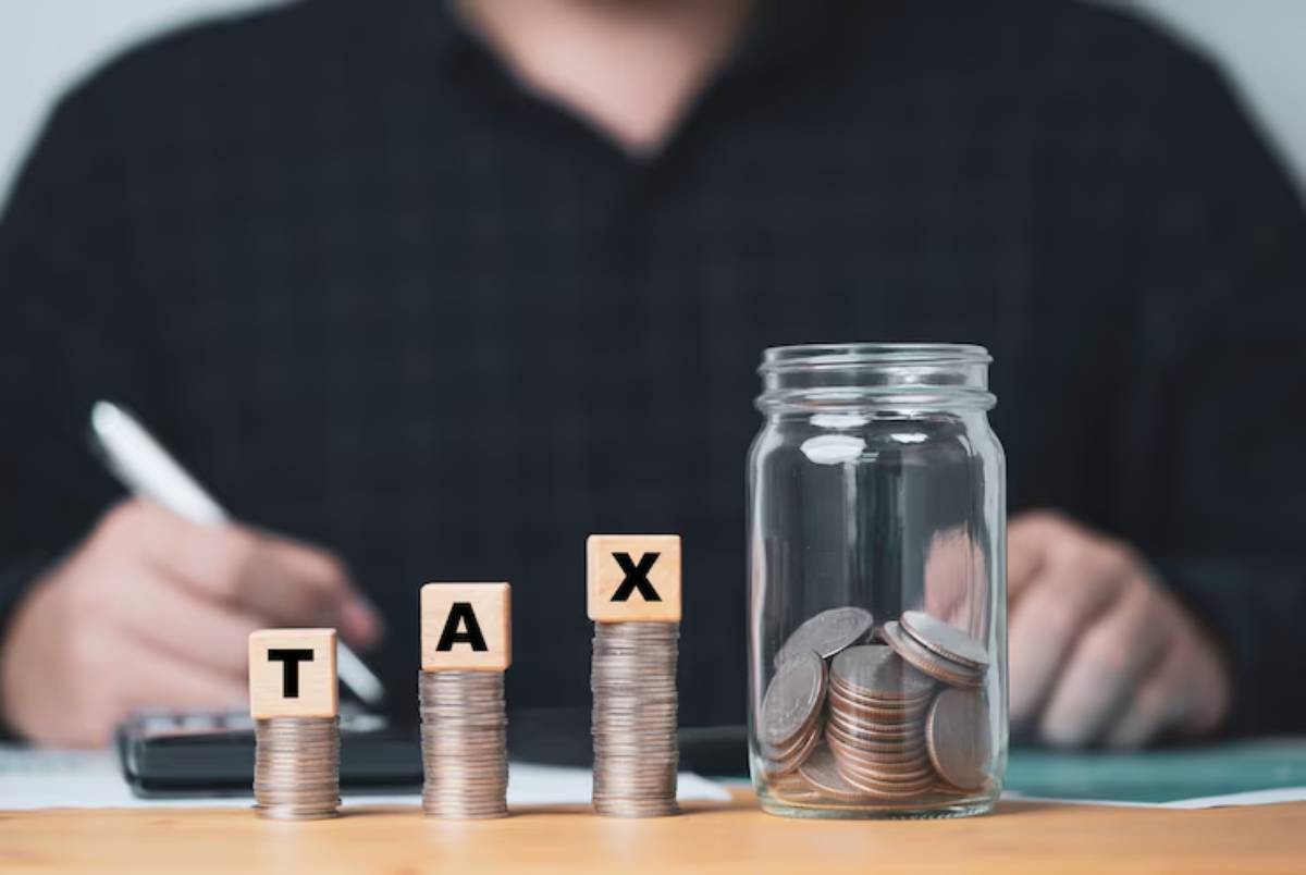 A person writes with a pen, surrounded by stacks of coins labeled TAX and a jar filled with coins, representing taxes.