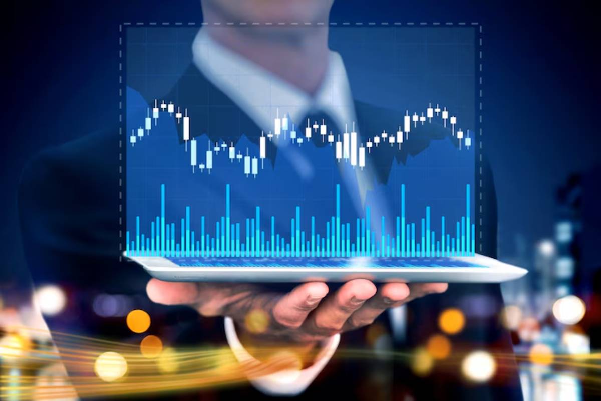 A professional in a suit holds a tablet displaying a financial graph with candlestick and bar charts against a blue, blurred background.