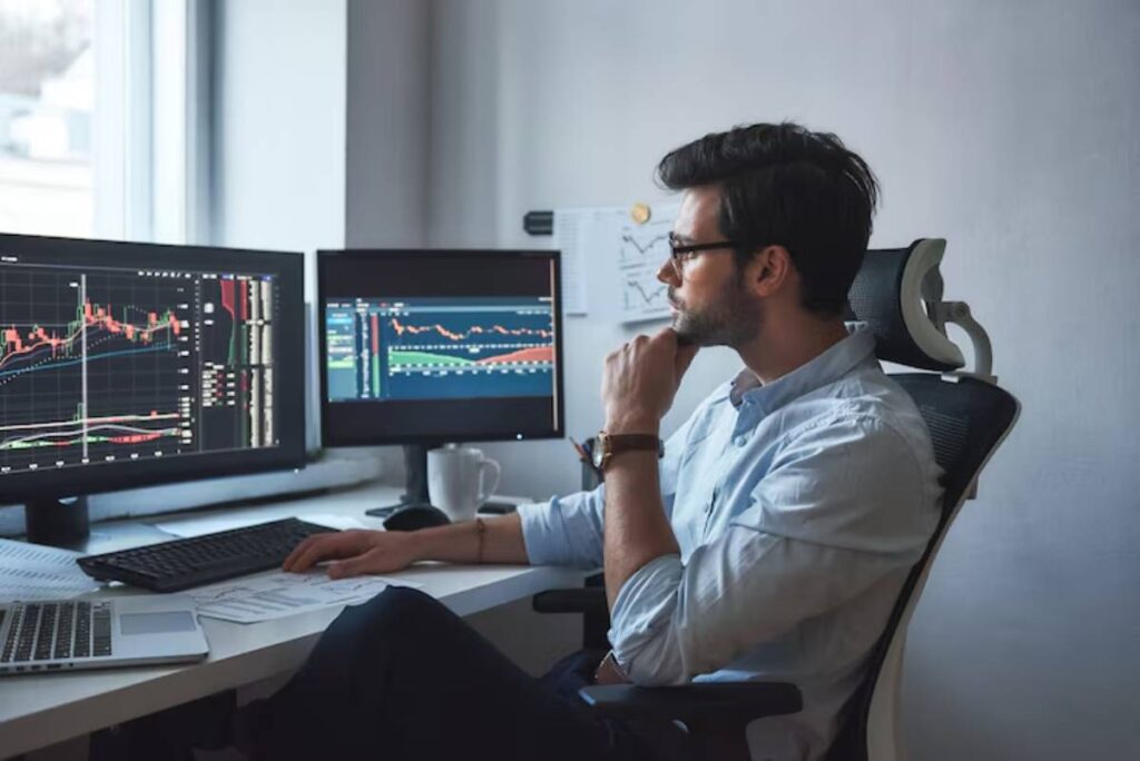 A man in a light blue shirt celebrates triumphantly at his dual computer setup, reviewing stock market graphs with glasses in hand.