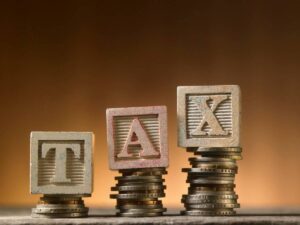 Wooden blocks spelling TAX balanced on stacks of coins, symbolizing finance and taxation concepts against a warm backdrop.
