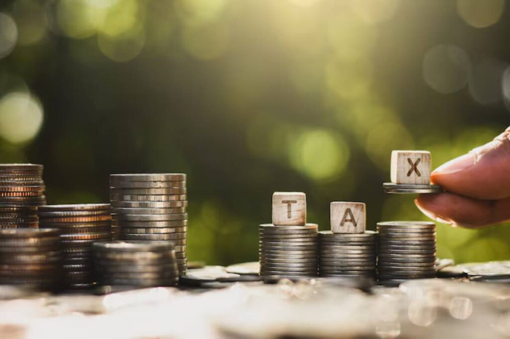 A hand places a wooden block labeled X atop stacks of coins, emphasizing tax financial concepts in a blurred background.