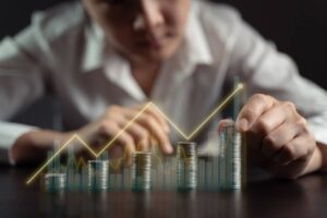 A person in a white shirt is arranging stacks of coins, with financial graphs and growth lines superimposed in the background.