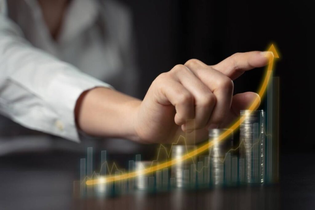 A hand points upward, signifying growth, above stacks of coins and a rising graph on a dark background.
