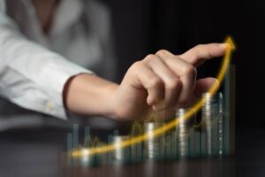 A hand points upward, signifying growth, above stacks of coins and a rising graph on a dark background.