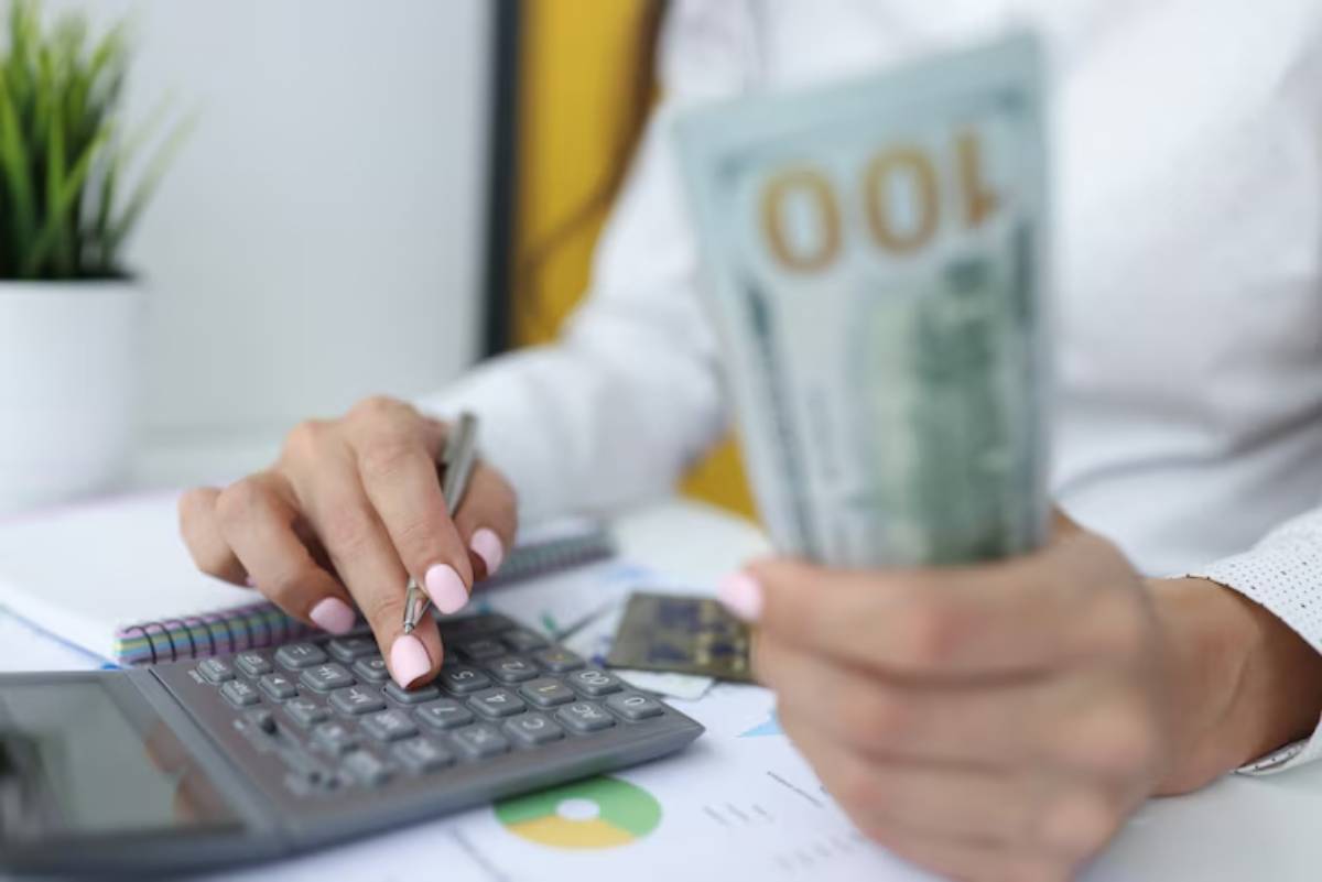 A person using a calculator, holding a $100 bill, with financial documents and a notebook on the desk.