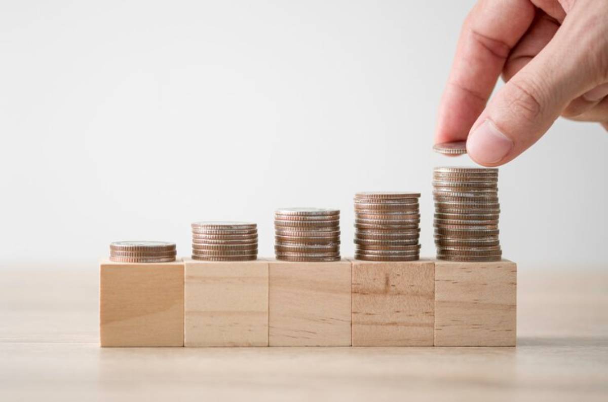 A hand stacks coins on ascending wooden blocks, illustrating growth or investment, with the background blurred for focus.