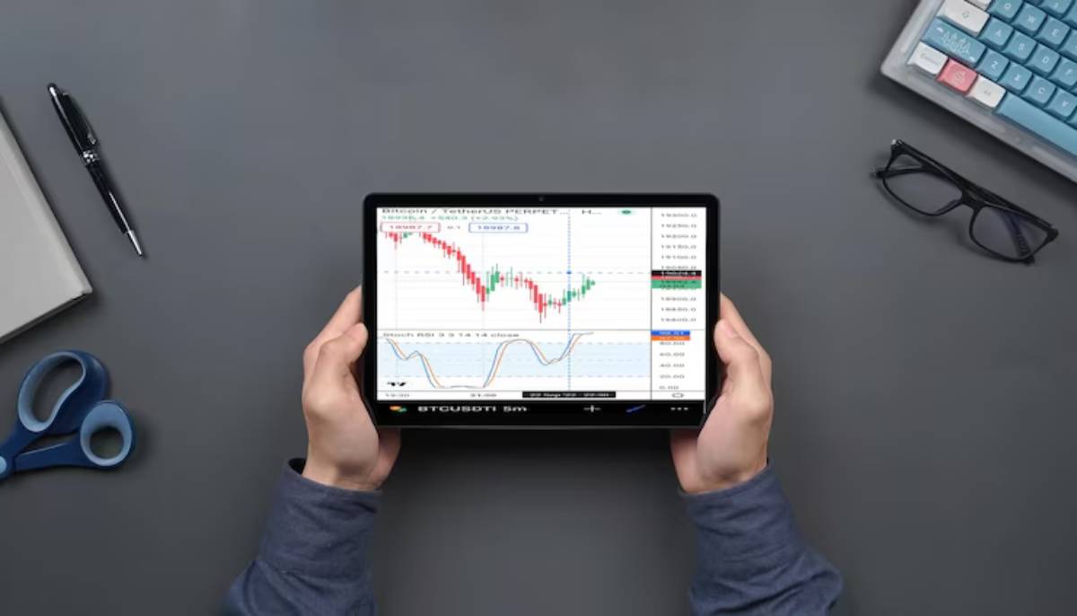 A person holds a tablet displaying Bitcoin trading charts, surrounded by a pen, notepad, scissors, and glasses on a desk.
