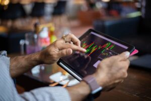 A person touches a tablet screen displaying colorful stock market graphs in a cozy, casual setting.