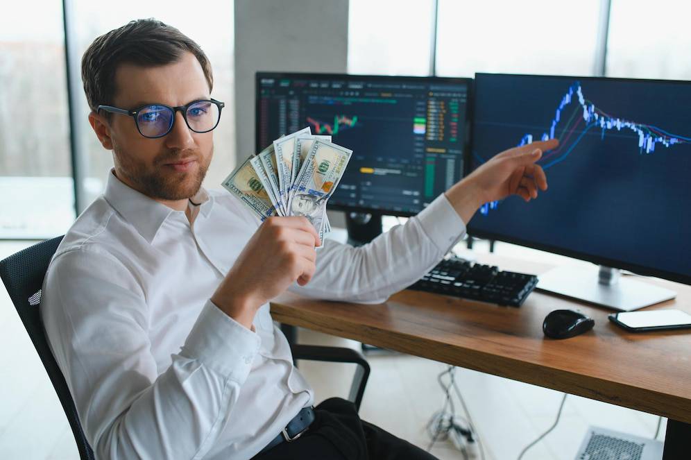 A person in a white shirt holds a fan of money while pointing at financial graphs displayed on dual monitors in a modern office setting.