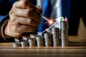 A hand holds a pen above stacks of coins, with an upward arrow and percentage signs, symbolizing financial growth and investment.
