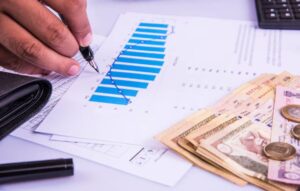 A hand holds a pen over financial documents showing a blue graph, while cash and coins are neatly arranged beside them.