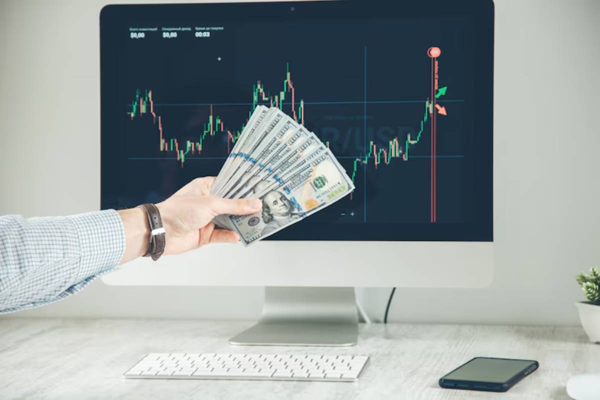 A person holds a bundle of cash in front of a computer displaying a fluctuating stock market graph.