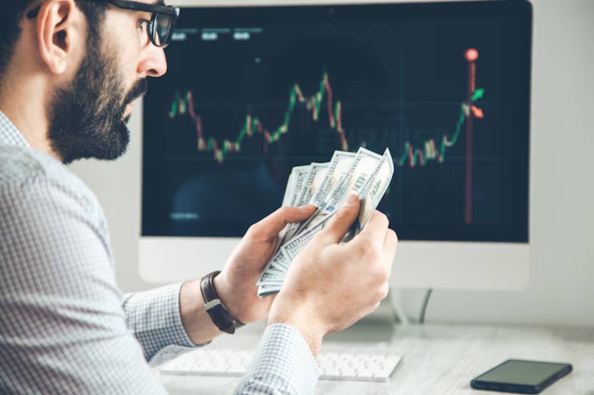 A person holds several banknotes while looking at a stock market chart on a computer screen, indicating a focus on trading.