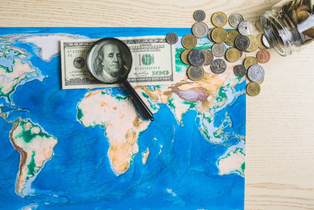 A magnifying glass rests on a hundred-dollar bill, surrounded by various coins, all atop a colorful world map.