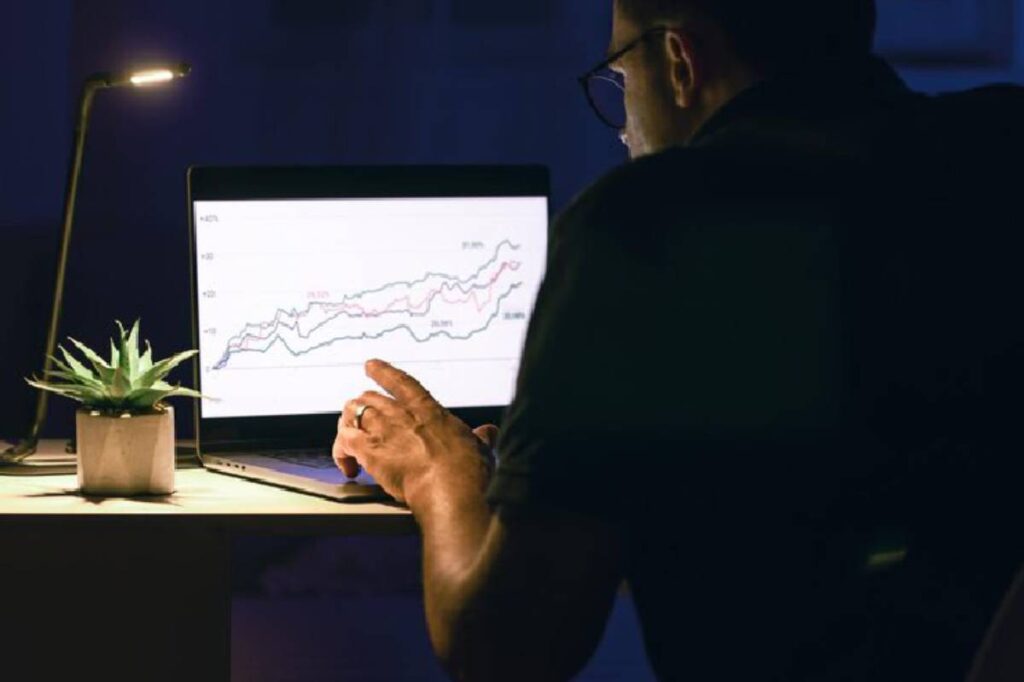 A person examines financial graphs on a laptop in a dimly lit room, with a small potted plant nearby.