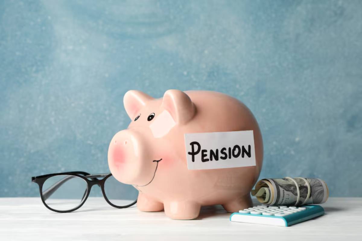 A pink piggy bank labeled PENSION, accompanied by glasses, rolled money, and a calculator on a light wooden table.