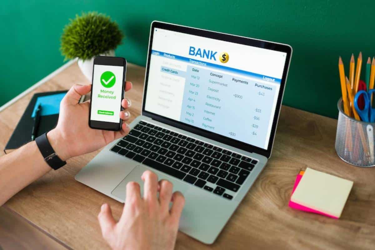 A person checks their phone displaying a Money Received notification while viewing bank transactions on a laptop.